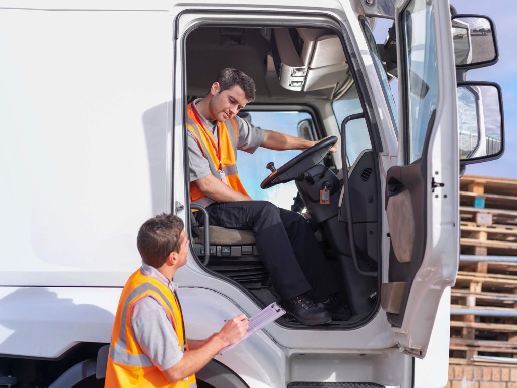 A lorry driver in his cab talking to a colleague holding a clipboard.
