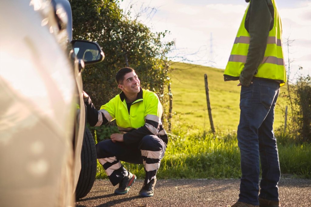 A technician crouched beside a car wheel at the roadside with a colleague in hi-vis.