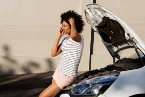 young woman standing by broken down car and making phone call